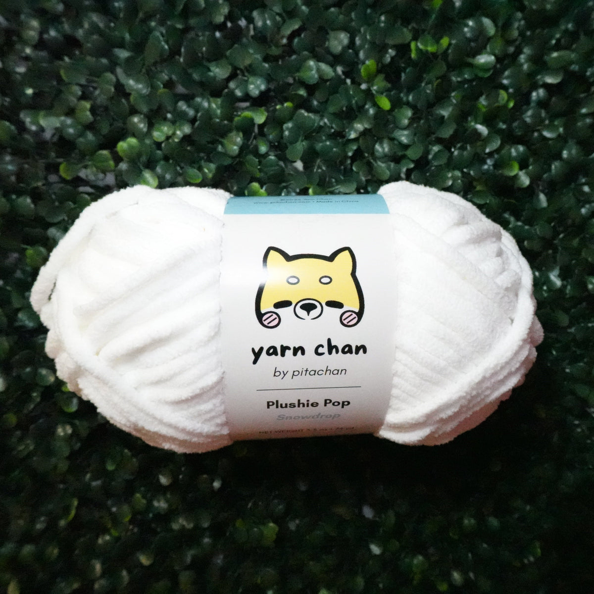 A skein of white yarn with a paper label reading 'yarn chan by pitachan' and 'Plushie Pop', placed on a dark green leafy textured background.